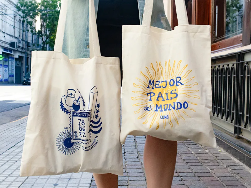 Dos tote bags de lienzo crudo estampadas en azul y amarillo, en una vereda empedrada de Buenos Aires